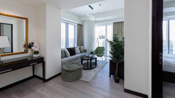 Una habitación de hotel moderna con sofá, una mesita redonda, un pequeño otoman, un escritorio con espejo y una planta en maceta.