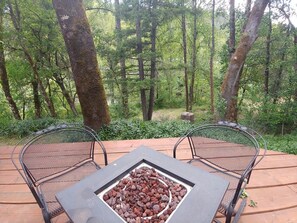 Ein Metalltisch und zwei Stühle auf einer Holzterrasse, umgeben von Bäumen.
