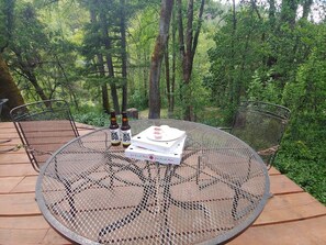 Ein runder Metalltisch mit einer Pizzaschachtel und zwei Flaschen mit dem Aufdruck "Joe's IPA" auf einer Holzterrasse, umgeben von Bäumen.
