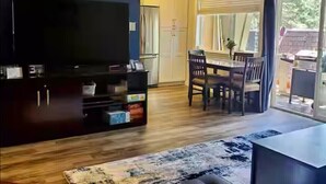 Een moderne woonkamer met een flatscreen televisie, donkere houten kasten, een eethoek met een tafel en stoelen, en een groot raam met gordijnen.