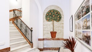 Un escalier avec une rampe en bois et des marches blanches, menant à un palier où se trouvent un arbre en pot et des photographies encadrées accrochées au mur.
