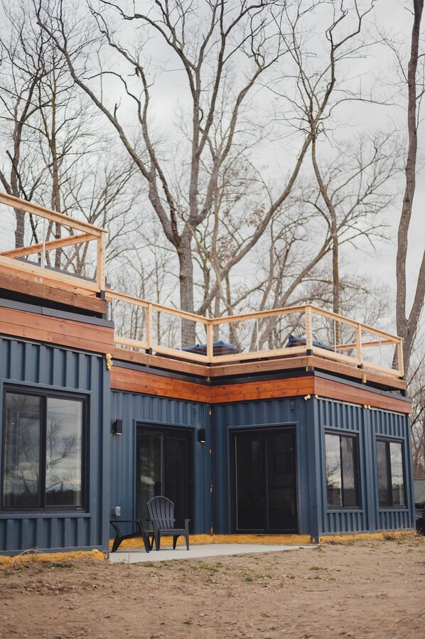 A modern building with a wooden deck on the upper level, large windows, and a chair outside. - Modern 4-bedroom container home on Long Lake! (Sarona)