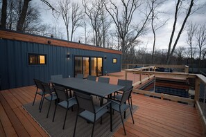 A modern outdoor deck with a dining table and chairs, a wooden deck, and a view of trees in the background. - Modern 4-bedroom container home on Long Lake! (Sarona)