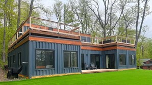 Una cabaña moderna de varios niveles con terraza de madera y ventanas amplias, rodeada de césped cuidado y árboles.