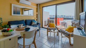 A modern living room with a sofa, dining table, and chairs, a TV, and a balcony with a view.