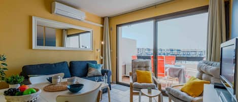 A modern living room with a sofa, dining table, and chairs, a TV, and a balcony with a view.