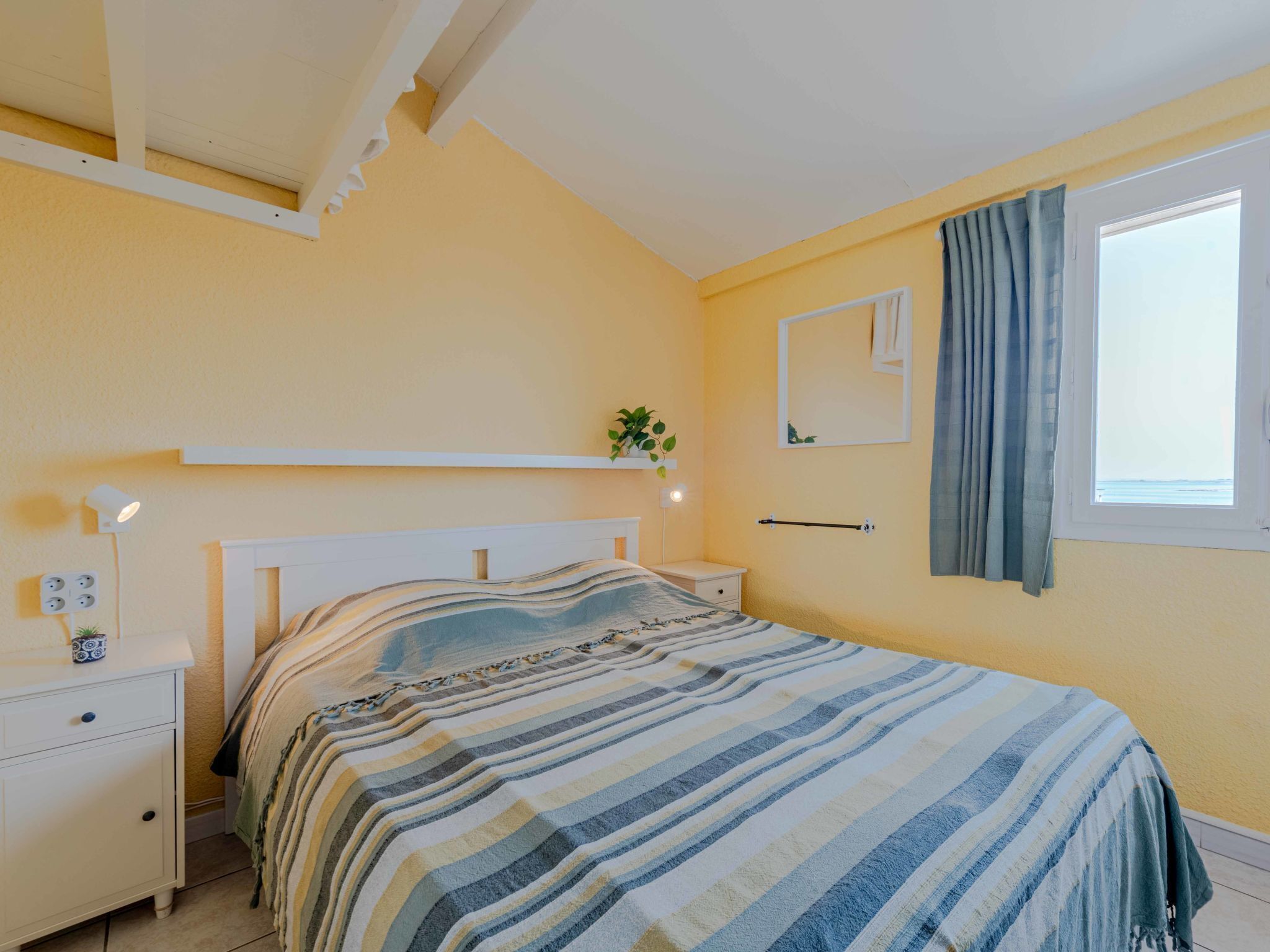 Un dormitorio con cama, mesitas de noche, un espejo, una ventana con cortinas y una planta pequeña en la pared.
