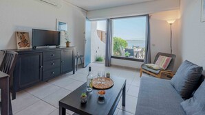 Una sala moderna con televisor de pantalla plana, una mesa de comedor con aperitivos, un sofá y un ventanal con vistas al mar.