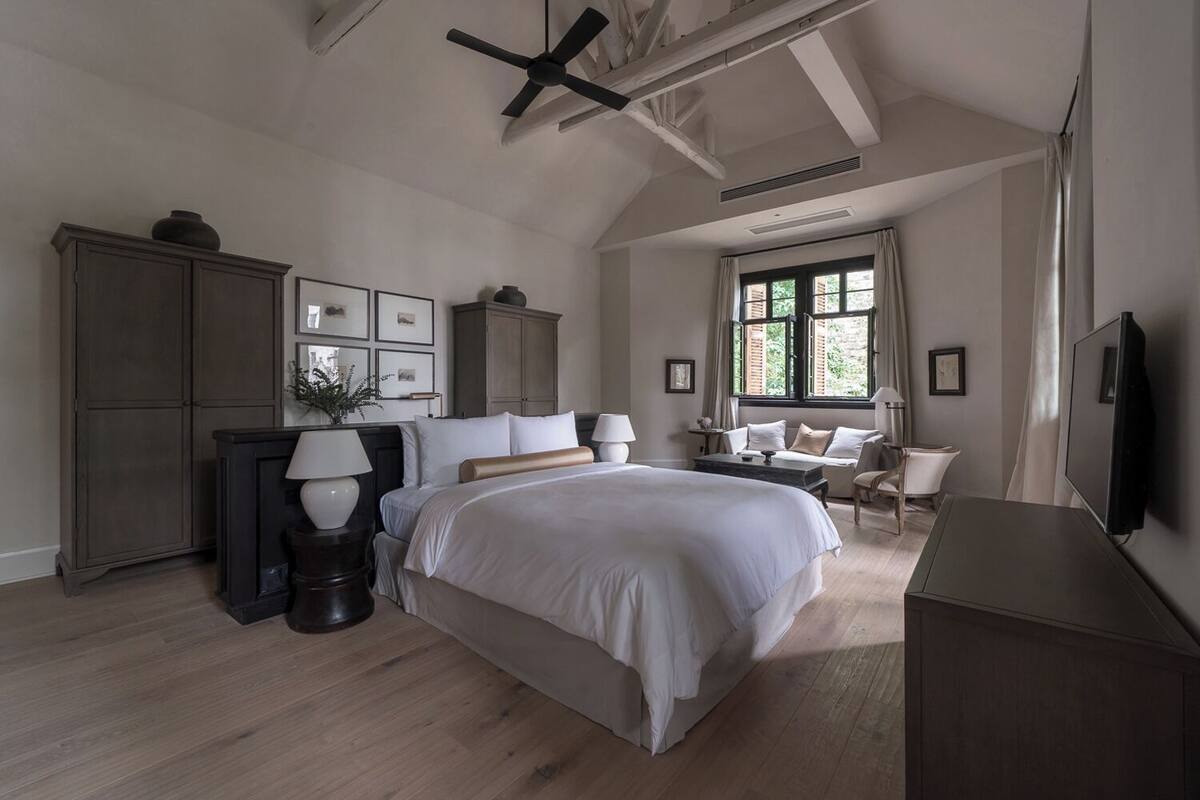 Une chambre avec un grand lit, un ventilateur de plafond, une télévision et une fenêtre avec des rideaux.