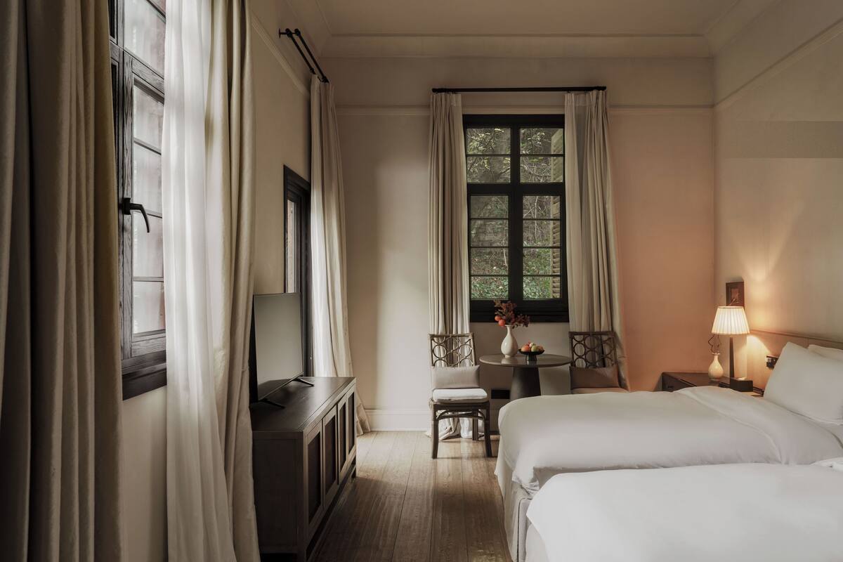Une chambre d’hôtel avec deux lits, un bureau, une chaise, une petite table avec un vase et une fenêtre avec des rideaux.