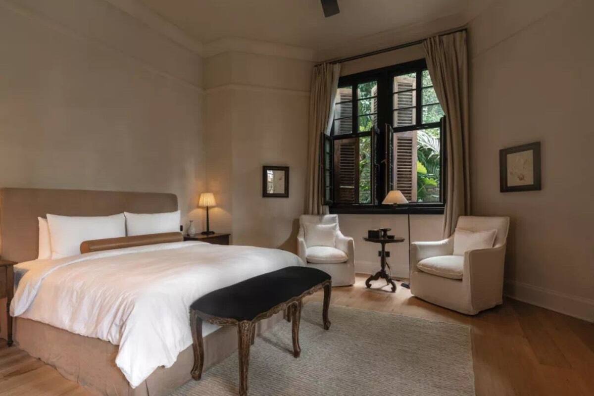 Une chambre à coucher avec un grand lit, deux fauteuils blancs, une petite table et une fenêtre avec des volets.