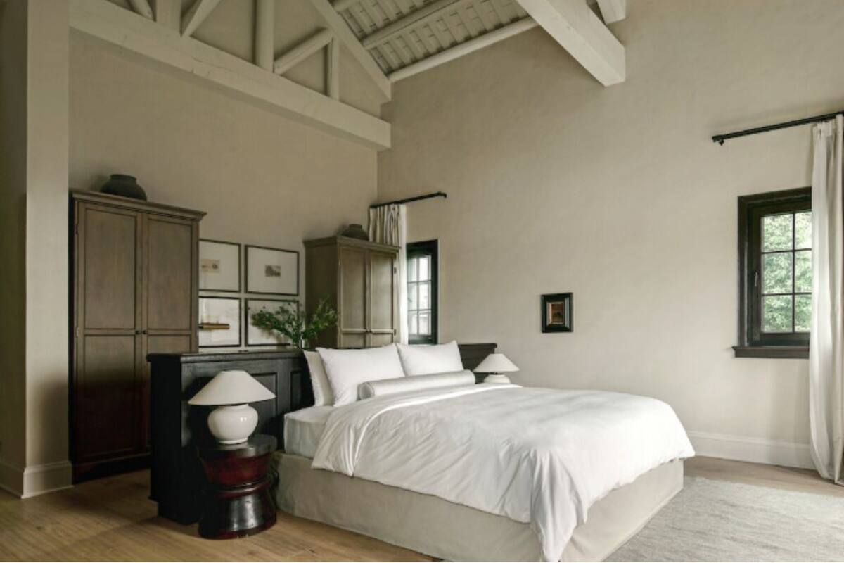 Une chambre à coucher avec un grand lit, une commode en bois foncé, une petite table avec une lampe et une fenêtre avec des rideaux.
