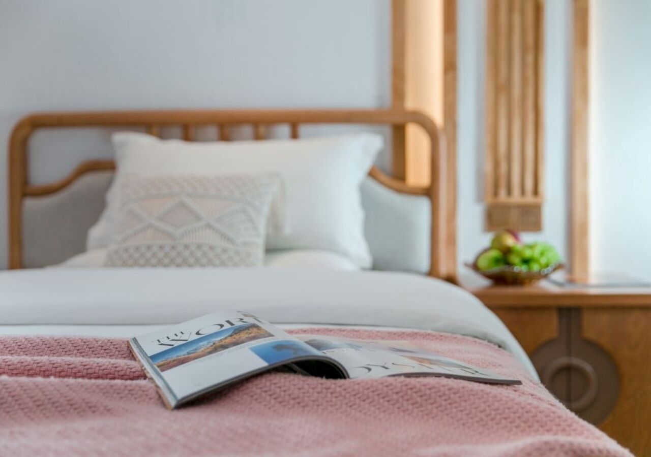 Cama bem arrumada com uma revista, cabeceira de madeira e mesa de cabeceira com uma cesta de frutas.