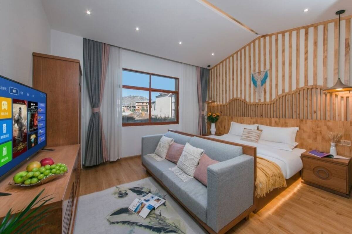 Una habitación de hotel moderna con pared de listones de madera, un ventanal con cortinas, un sofá gris, cama con ropa de cama blanca y una mesita de noche de madera.