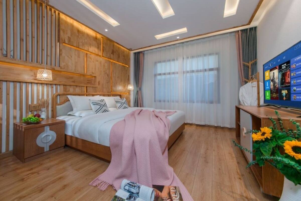 Un dormitorio moderno con cabecera de madera, cama con manta rosa, mesita de noche con lámpara y televisor sobre un mueble de madera.