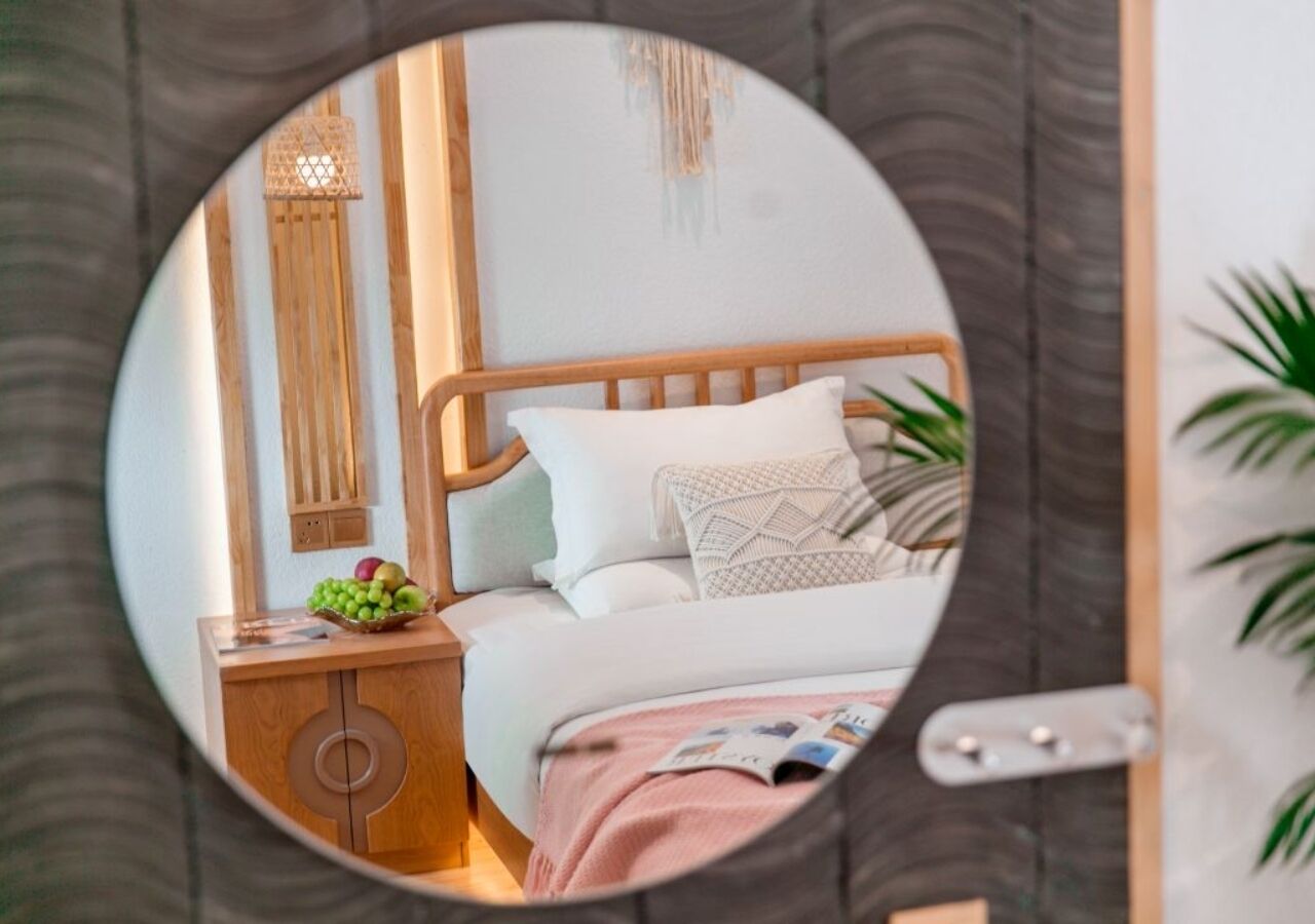 A bedroom with a wooden headboard, a bedside table with a fruit bowl, a magazine on the bed, and a mirror reflecting the room.