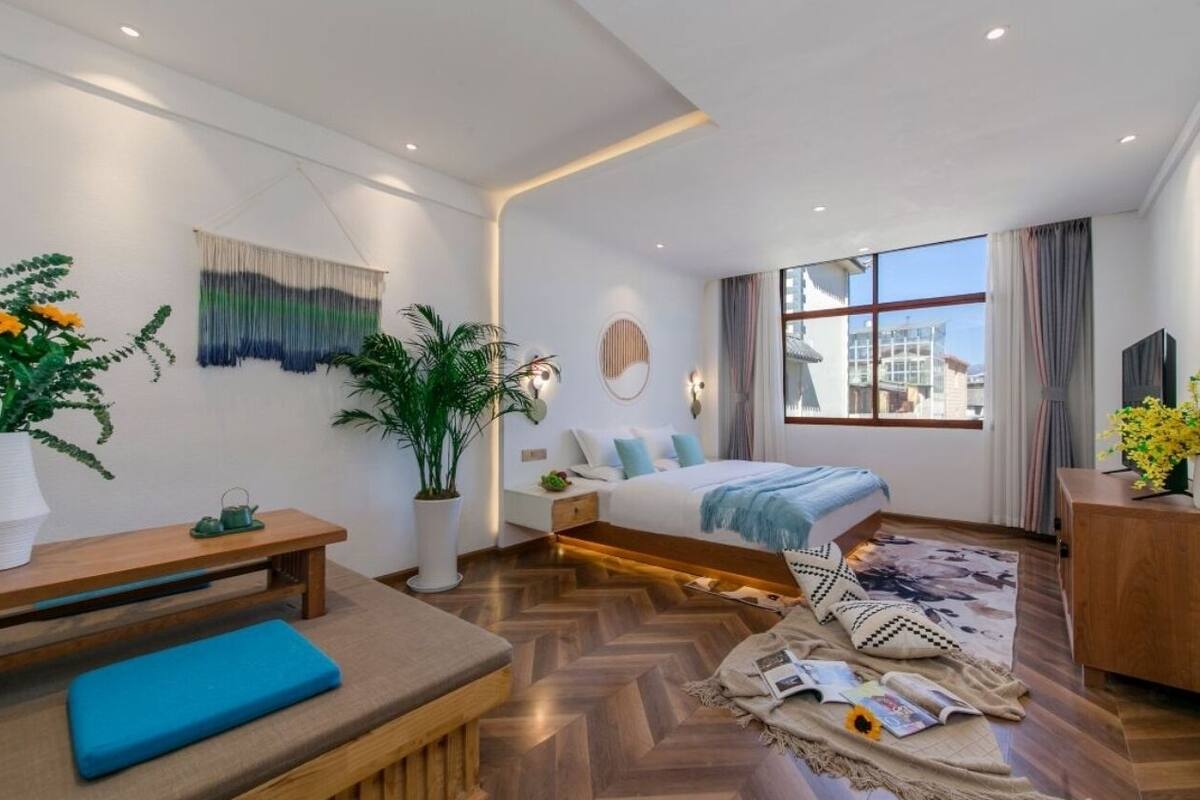 Una habitación de hotel moderna con una cama grande, una zona de estar con un banco, una mesita y un ventanal con vistas a la ciudad.