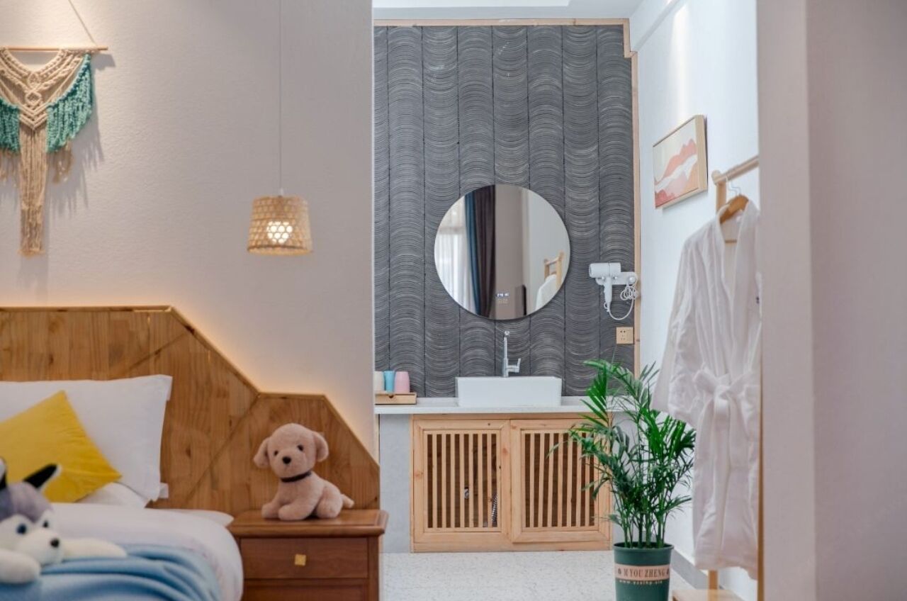 A bedroom with a wooden headboard, a bed with a teddy bear, a nightstand, a bedside lamp, a mirror, a sink, a cabinet, and a potted plant.