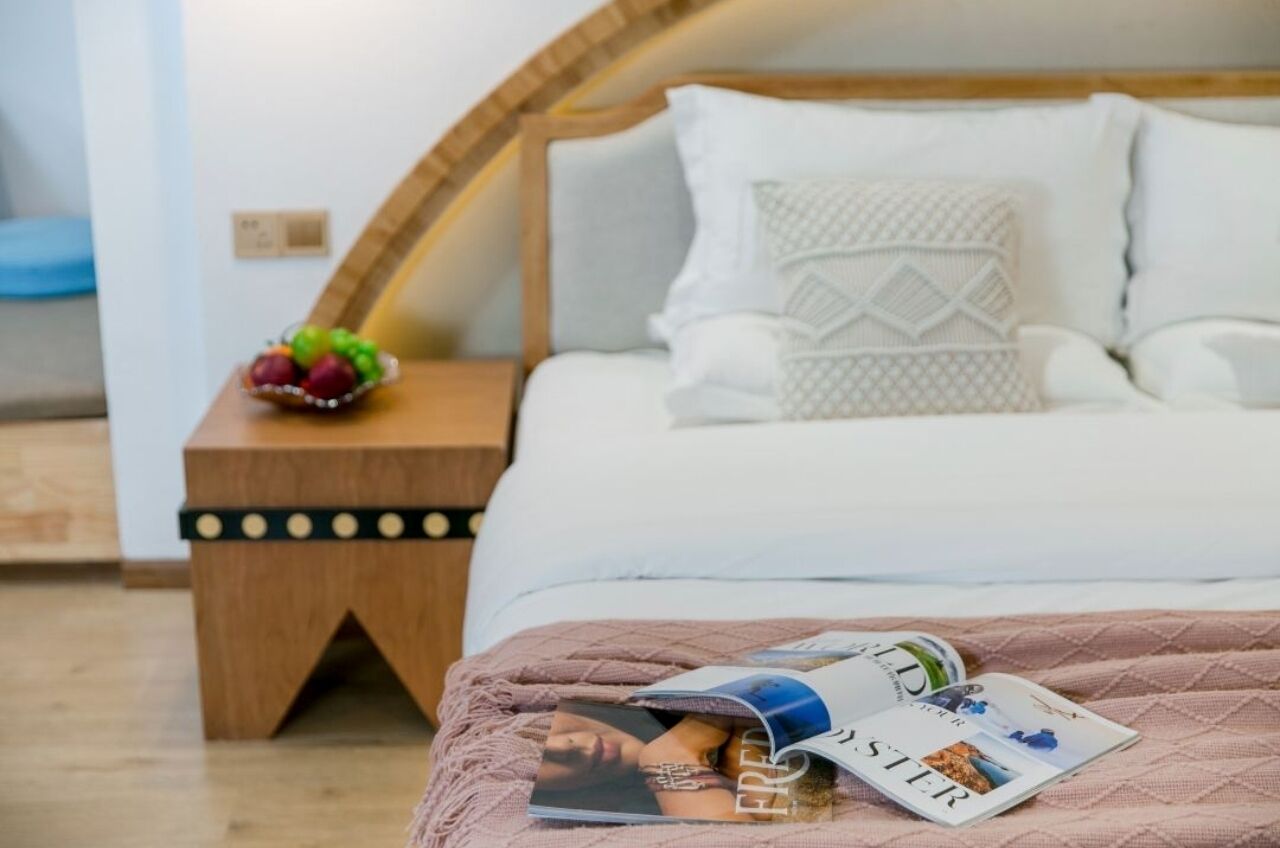 Ein ordentlich bezogenes Bett mit einer rosa Decke, ein Nachttisch aus Holz mit einer Obstschale und eine Zeitschrift auf dem Bett.