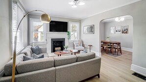 A living room with a grey sofa, a fireplace, a TV, and a dining area with a table and chairs. - Hot Tub + Big Yard • Minutes from Uptown! (Charlotte)