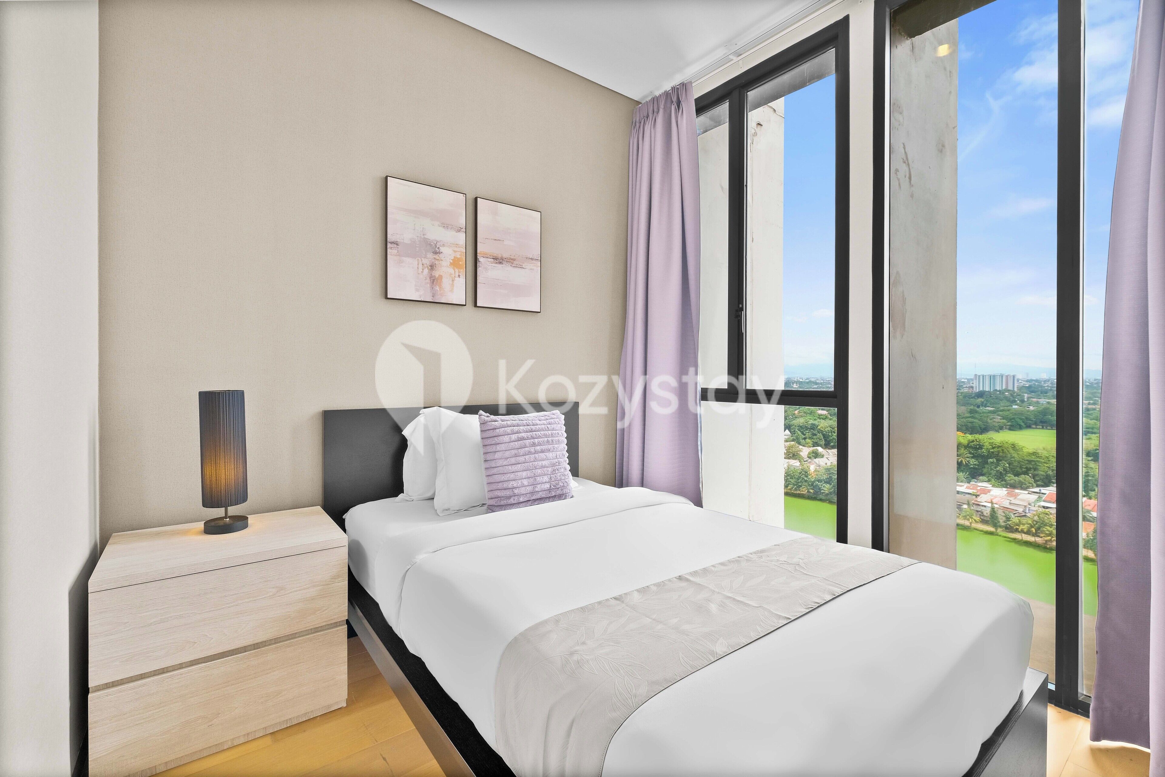 Ein modernes Schlafzimmer mit einem großen Bett, einem Nachttisch, einer Lampe und Blick auf eine Stadtkulisse durch große Fenster.