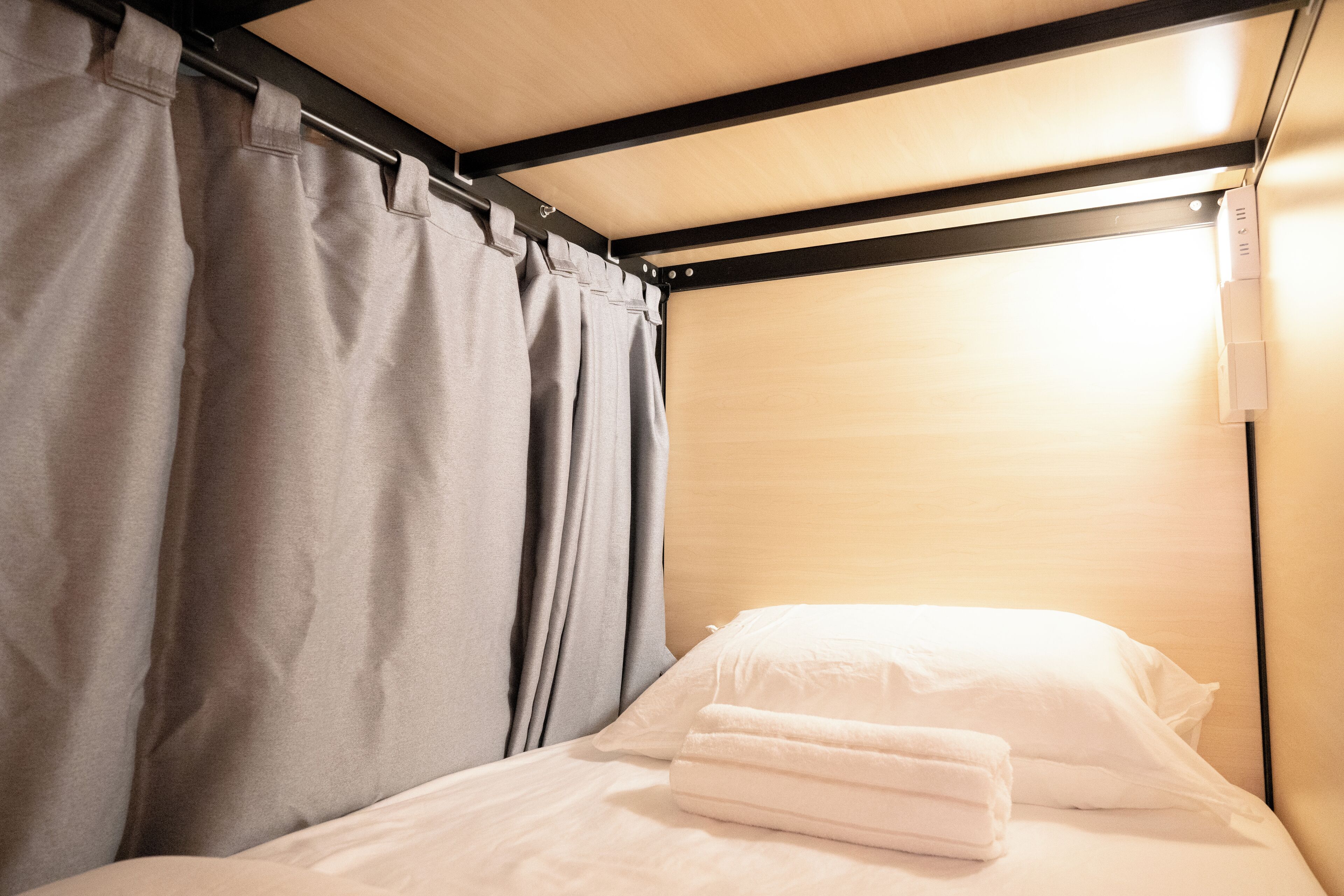 A single bed with white linens and a grey curtain, a wooden headboard, and a light fixture above.