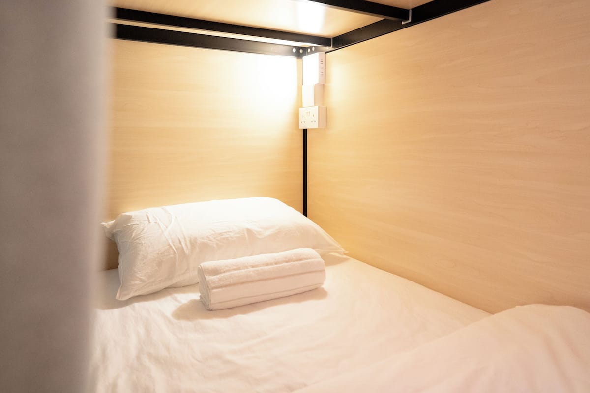 A small, neatly made bed with white linens and a rolled towel, a wall socket, and a wooden panel wall.