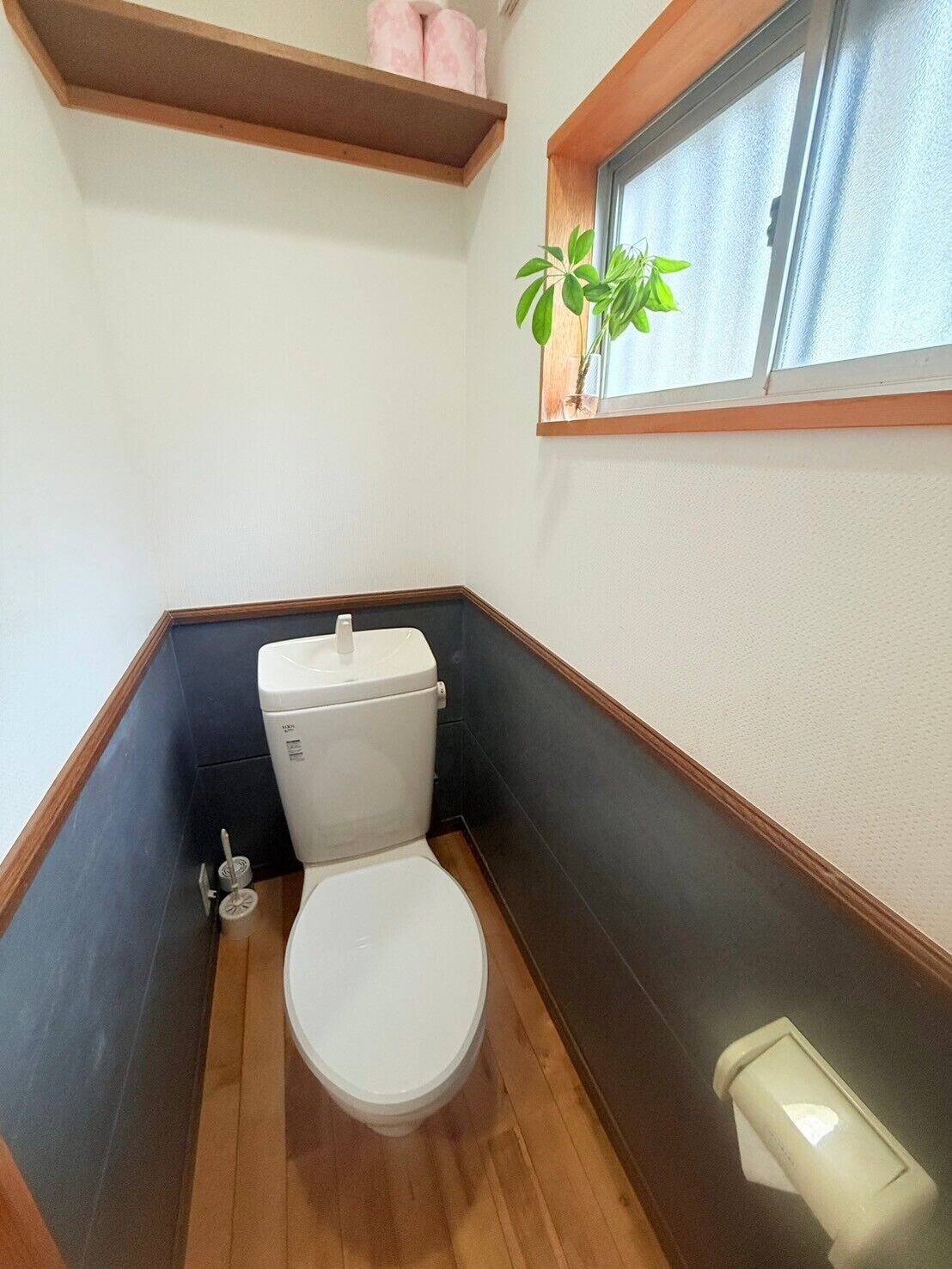 A small, compact bathroom with a white toilet, a wooden floor, a window with a plant, and a shelf with towels.
