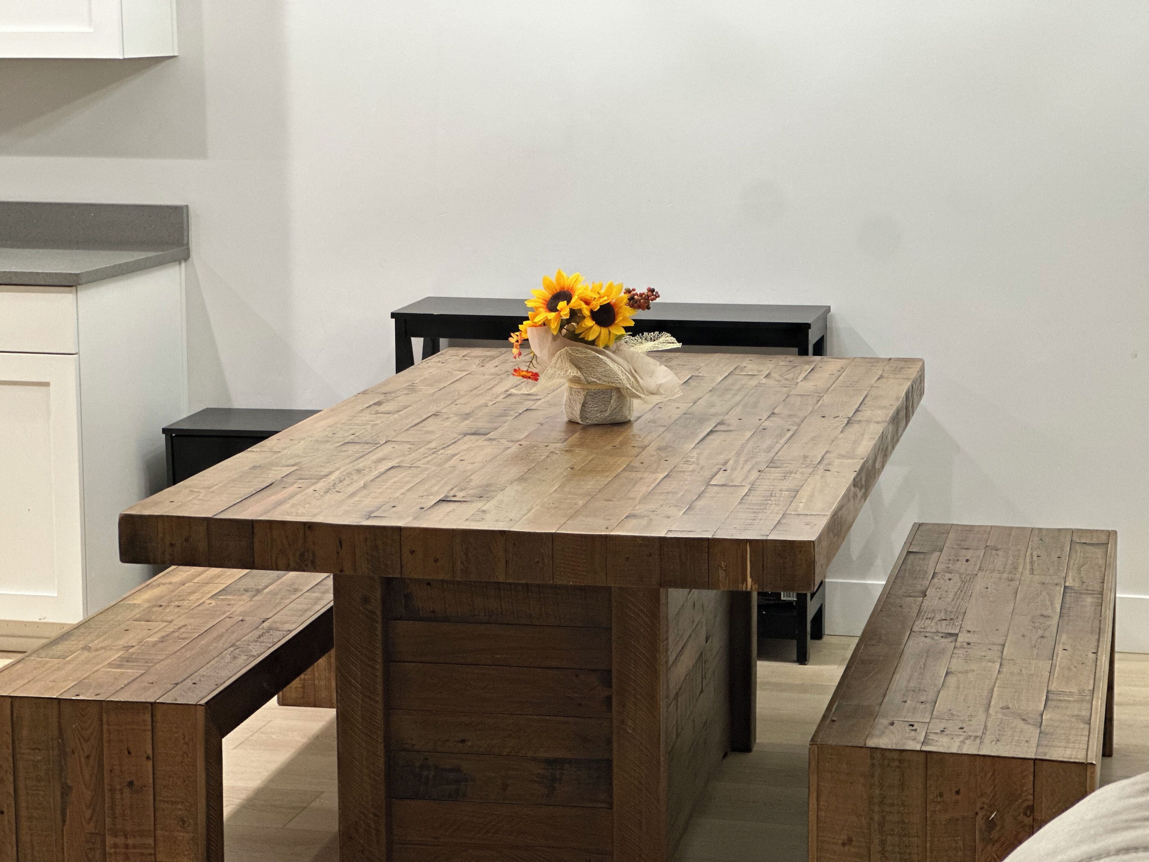 Ein Holzespeisetisch mit Bänken, eine Vase mit Sonnenblumen und ein schwarzer Tisch an einer weißen Wand.