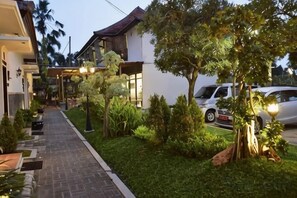 Un camino que lleva a un edificio con un área de estar cubierta al aire libre, rodeado de vegetación y coches aparcados.