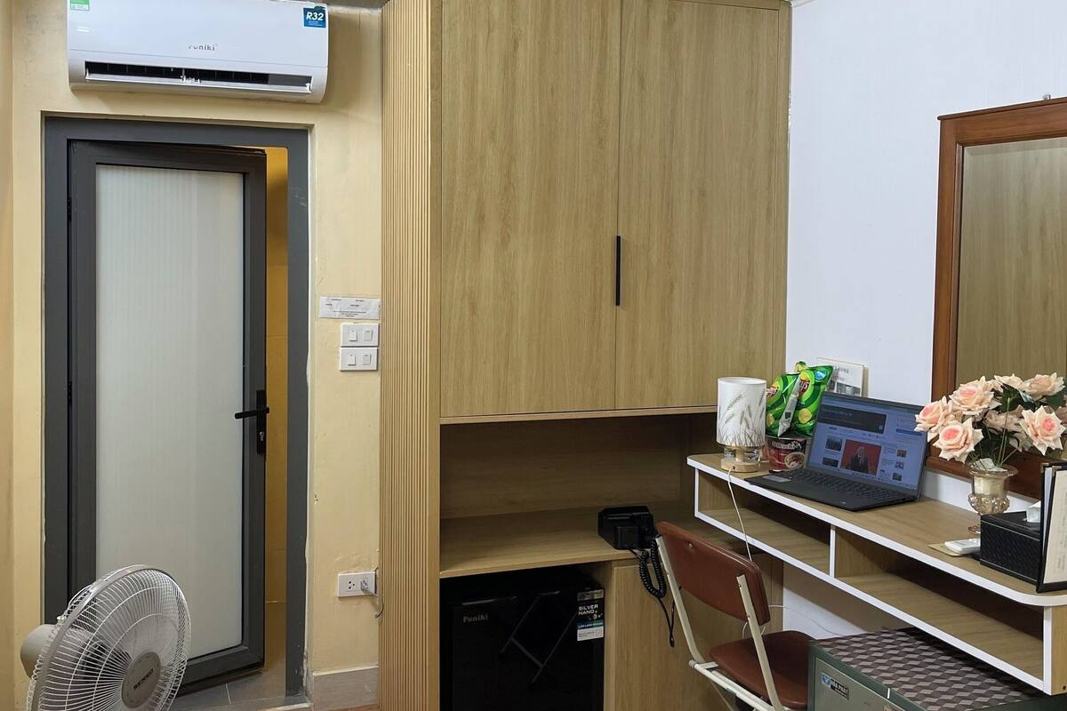 A compact room with a wooden wardrobe, a desk with a laptop, a small fan, and a safe.