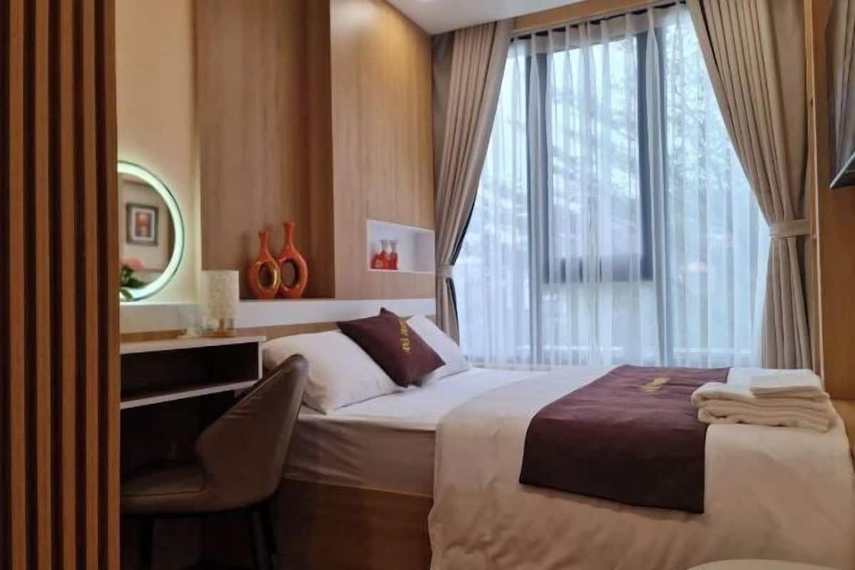 A hotel room with a bed, a desk, a chair, a small shelf with decorative items, and a window with curtains.