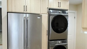 Une cuisine équipée d’appareils électroménagers en acier inoxydable, notamment un réfrigérateur et un appareil combinant lave-linge et sèche-linge, avec des meubles de couleur bois clair et un plan de travail blanc.
