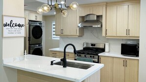 Une cuisine moderne dotée d’un îlot central, d’appareils électroménagers en acier inoxydable, de meubles de couleur bois clair et d’un luminaire décoratif.