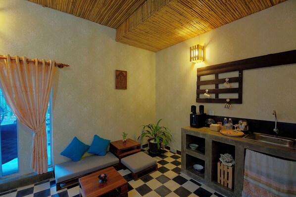 A cozy room with a checkered floor, a sofa with blue cushions, a small table, a potted plant, and a kitchenette with a sink and shelves. - Old Khmer House (Siem Reap)