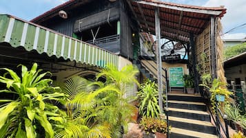 Un bâtiment traditionnel en bois, doté d’un escalier menant à une zone abritée, le tout entouré d’une végétation luxuriante.