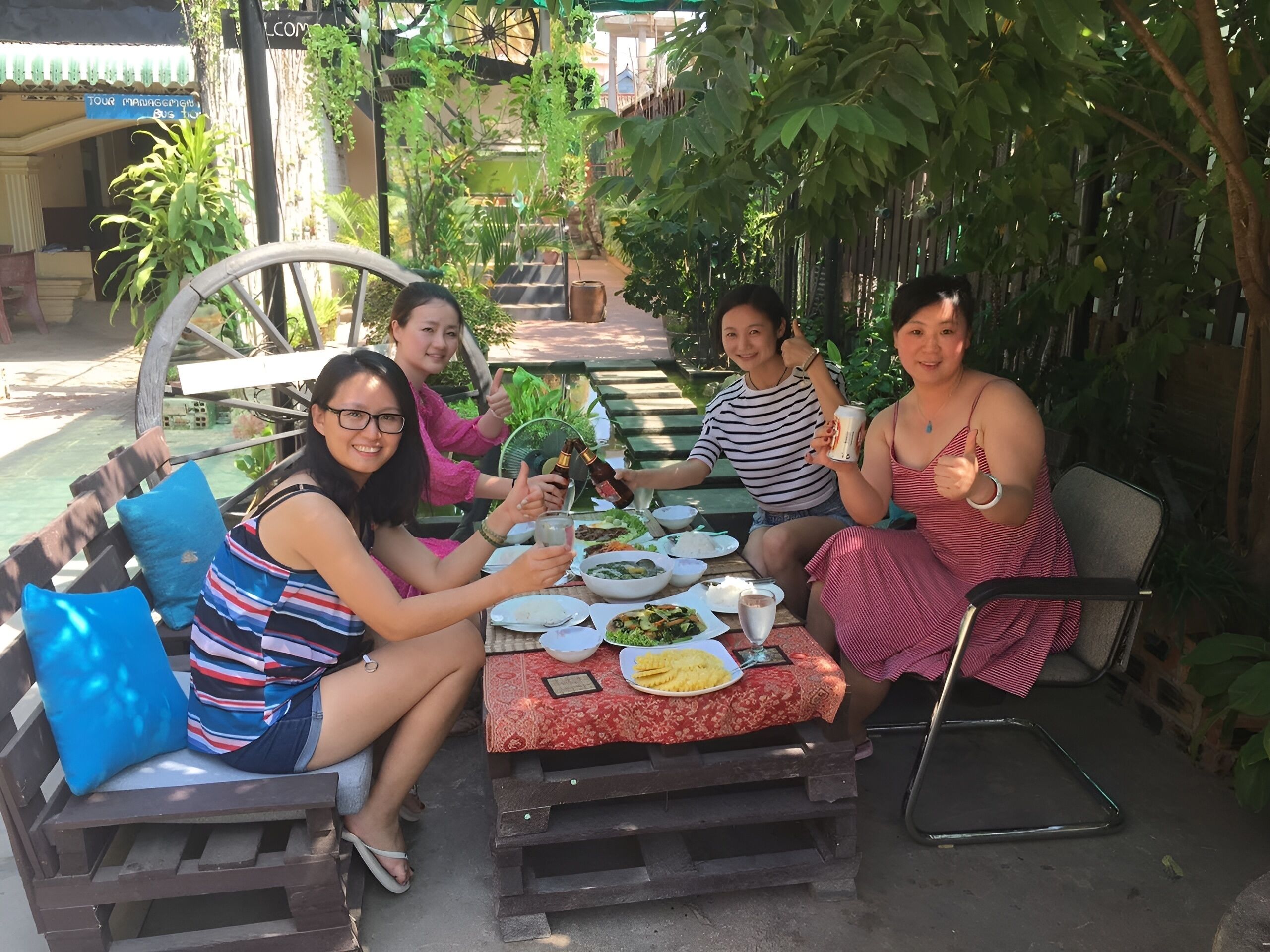 A group of people sitting at an outdoor table with various dishes, surrounded by greenery and a wooden bench.