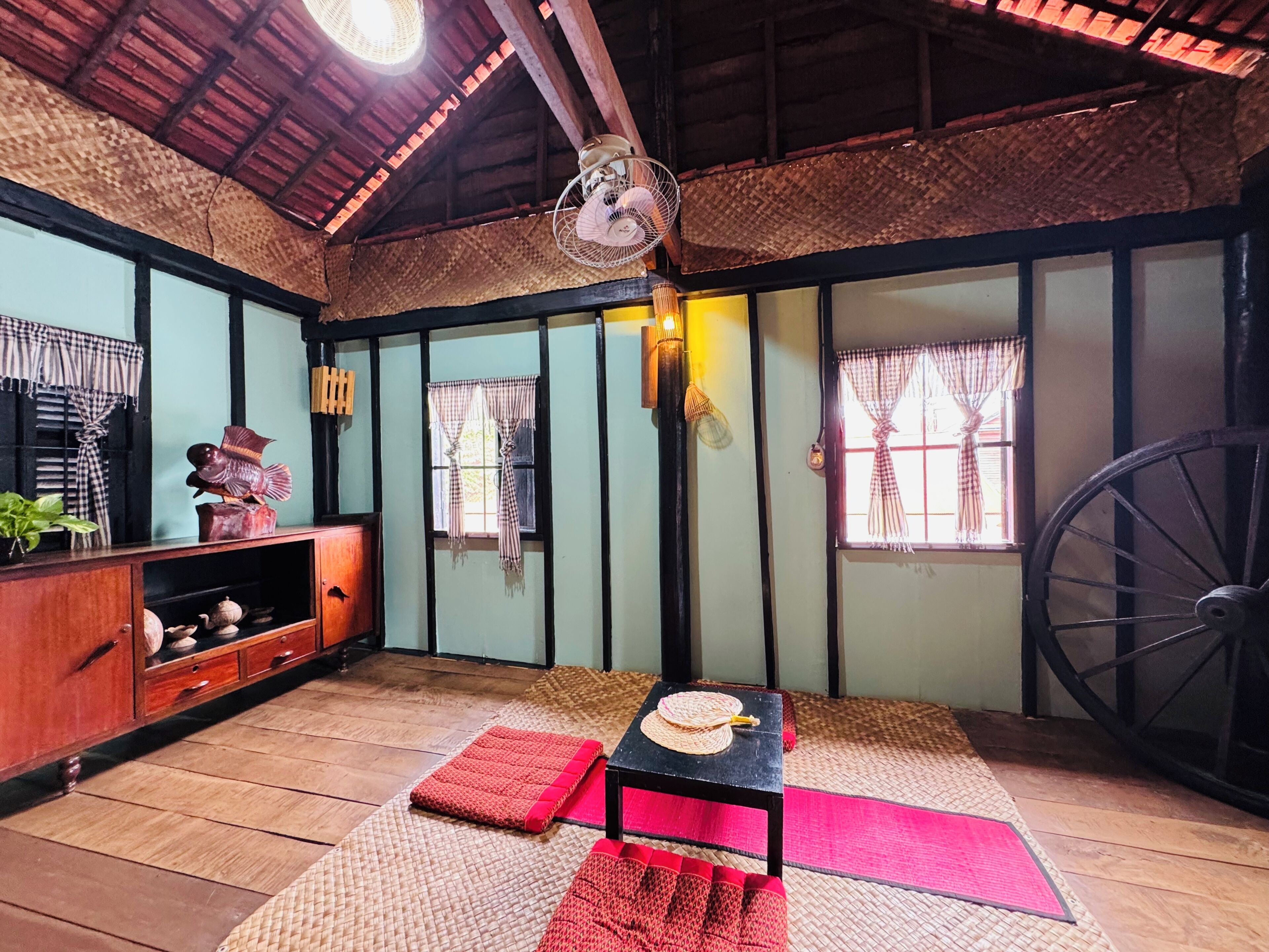 A traditional room with a wooden floor, a black table with a woven mat, a large wheel, and a fan.