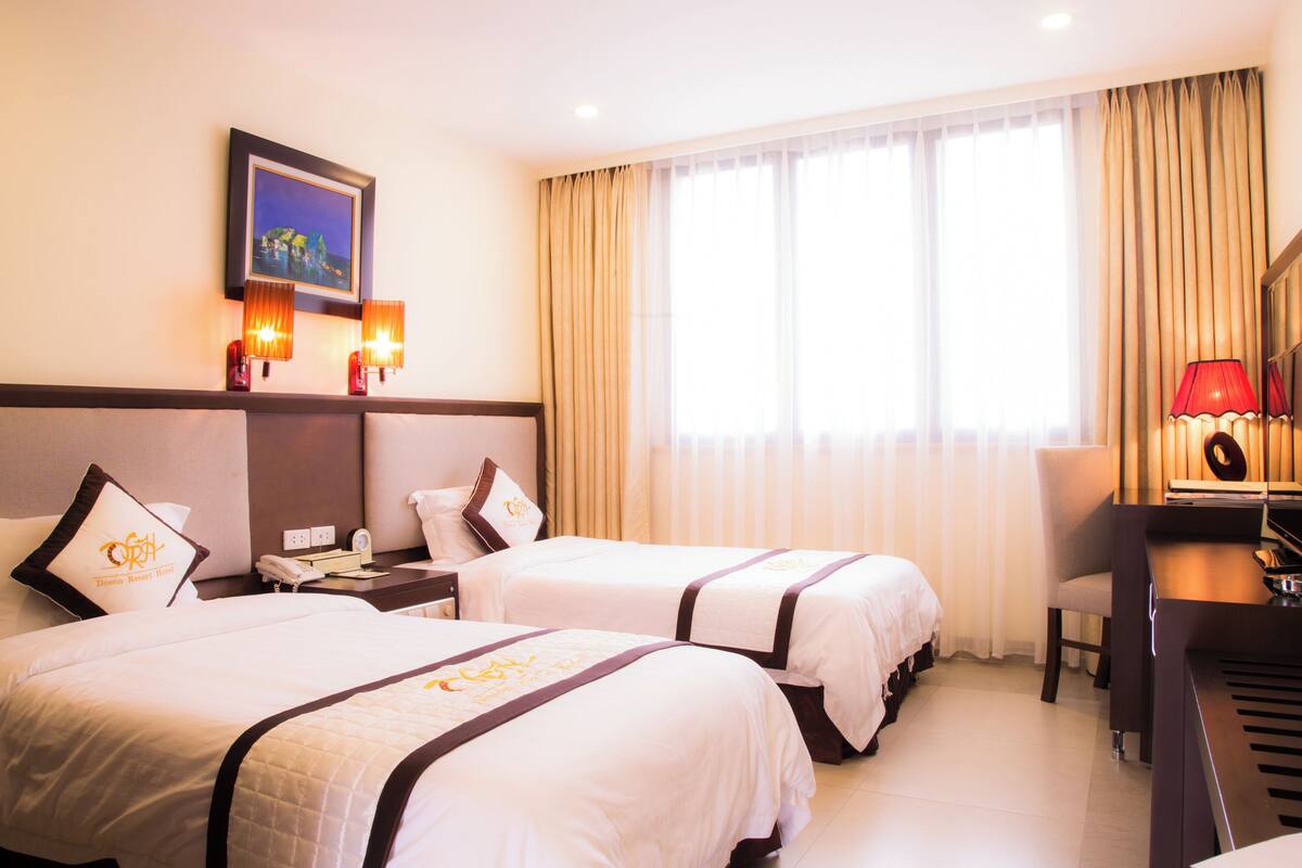 A hotel room with two beds, a desk with a piano, a chair, a nightstand with a lamp, and a window with curtains.
