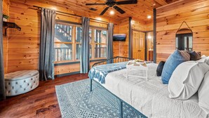 A wooden cabin bedroom with a bed, a ceiling fan, a window with curtains, and a small round ottoman. - Sleeps 32: Perfectly Designed For Groups/Reunions! (Gatlinburg)