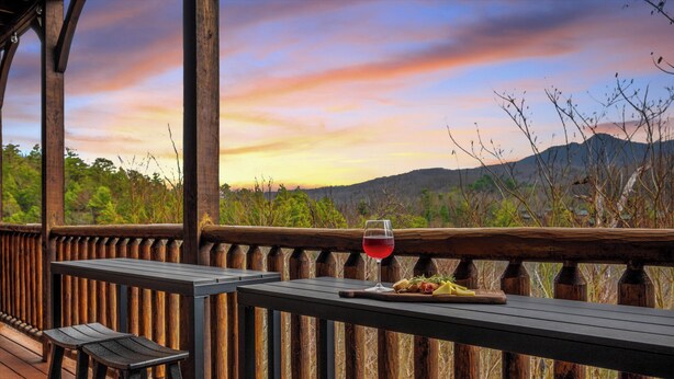 A wooden deck with a table and two stools, a glass of red wine, and a plate of food. - Sleeps 32: Perfectly Designed For Groups/Reunions! (Gatlinburg)