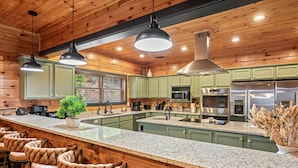 A spacious kitchen with granite countertops, green cabinets, and stainless steel appliances. There are bar stools along the counter and a potted plant on the island. - Sleeps 32: Perfectly Designed For Groups/Reunions! (Gatlinburg)