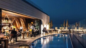 Un espace aménagé au bord de la piscine, comprenant un bar et un coin repas, ainsi qu’une piscine et une façade de bâtiment au design moderne.