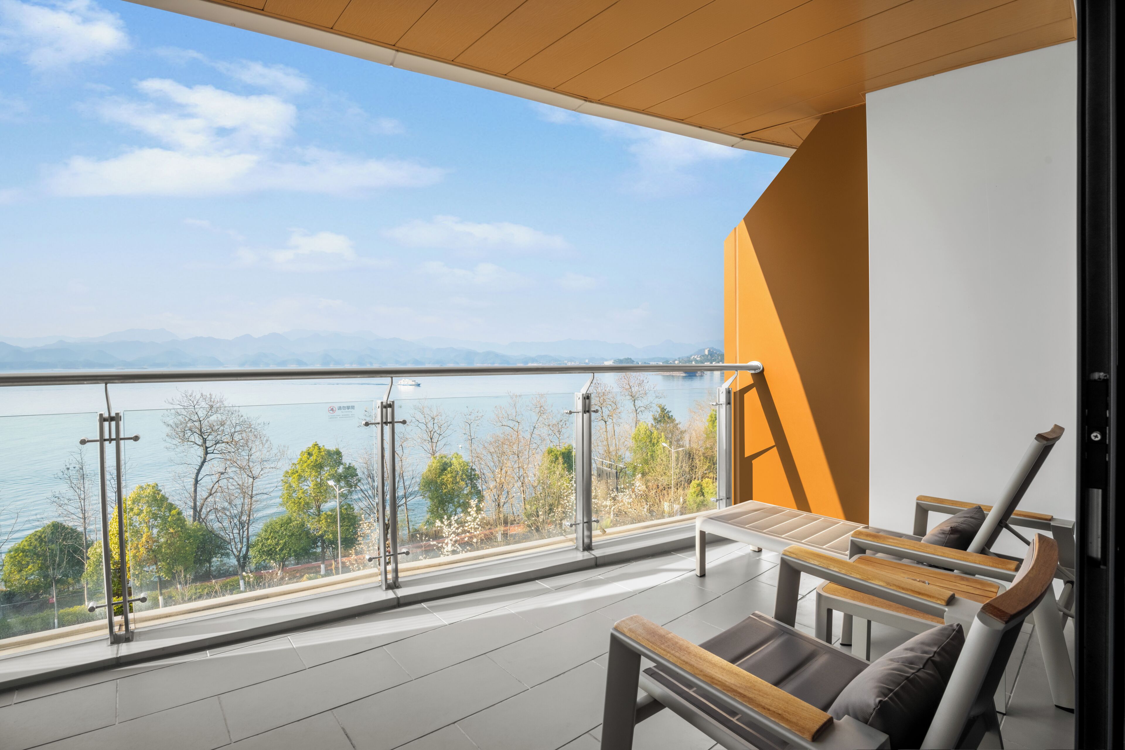 Un balcón con vistas al agua y montañas, amueblado con sillas y una mesa de madera.