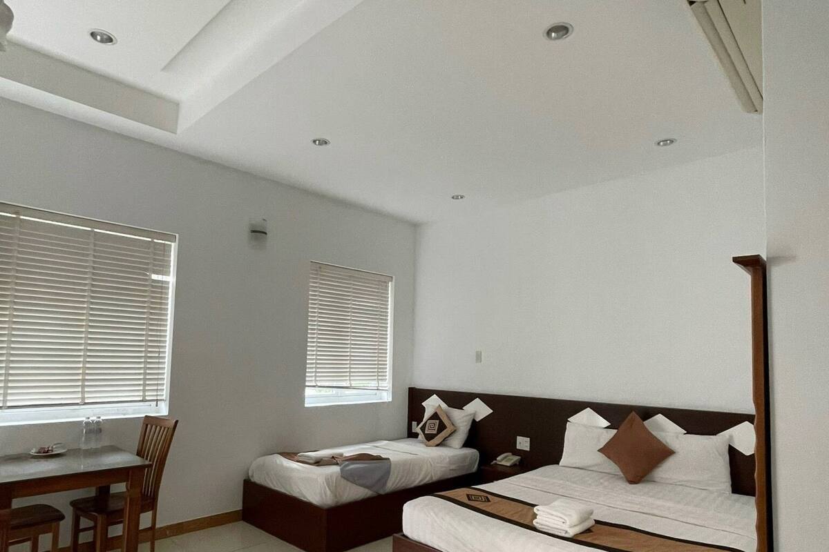 A modern hotel room with a dining area, two single beds, a desk, and a large window.