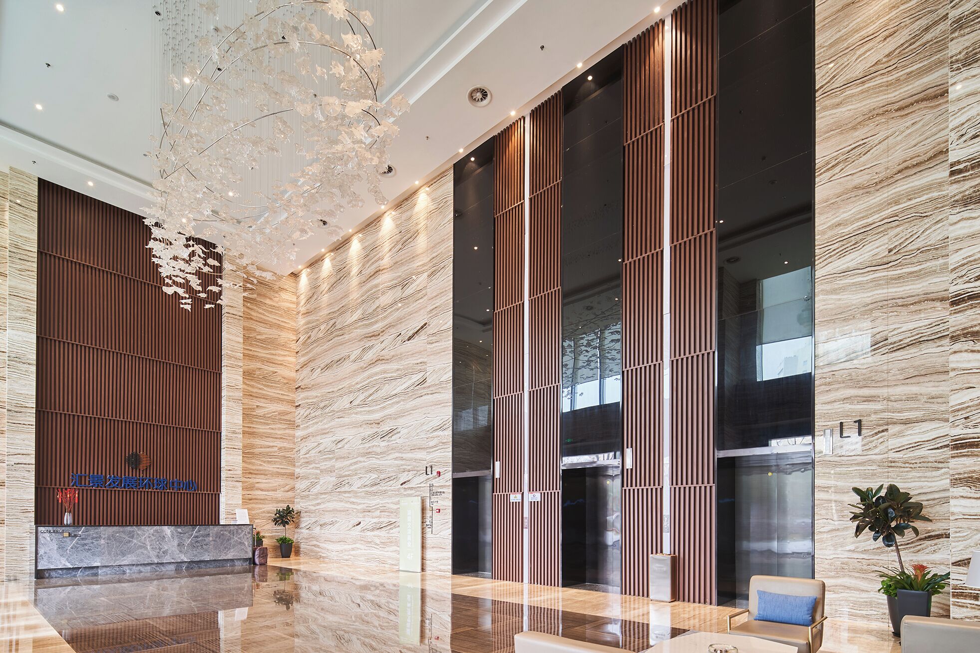 Een moderne lobby met een marmeren vloer, een grote kroonluchter, verticale houten lamellen en glazen liften.