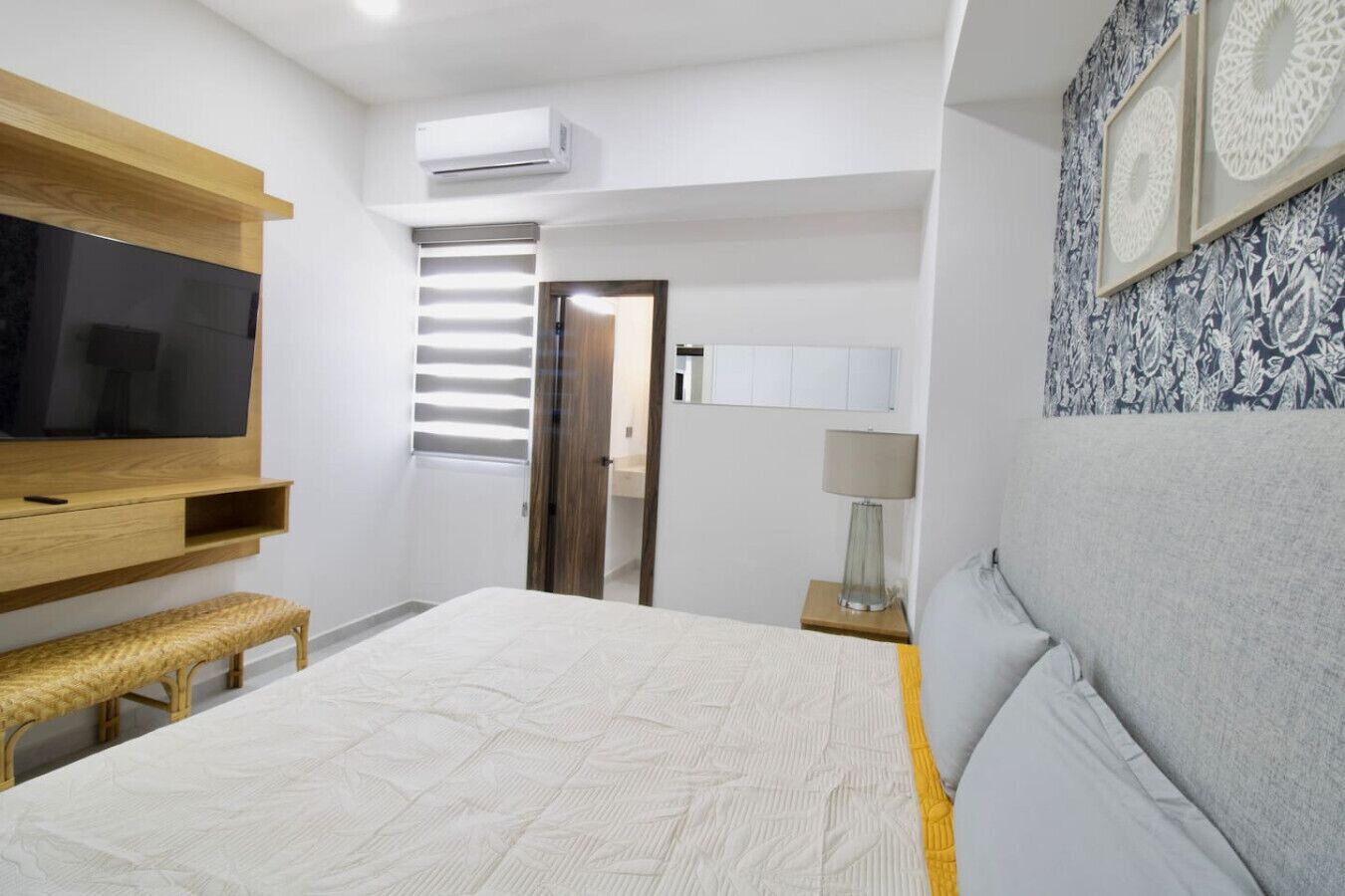 A modern bedroom with a large bed, a TV mounted on the wall, a bench, a bedside table with a lamp, and a patterned wall.