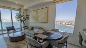 A modern living room with a round table, grey sofas, and a large window offering a city view.