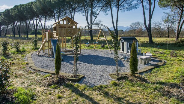 Un’area giochi con una casetta di legno, altalene e uno scivolo, circondata da alberi e un vialetto sterrato.