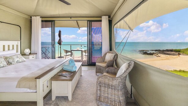 A bedroom with a bed, a bench, and a wicker chair, overlooking a beach with a view of the ocean and a boat.